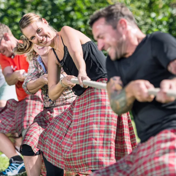 Gruppe Menschen im Kilt zieht lachend an einem Tau im Freien.