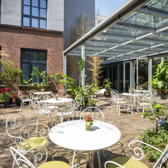 Café-Terrasse mit weißen Tischen, Stühlen und vielen Pflanzen im Grünen.