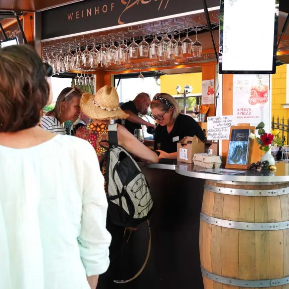 Menschen stehen an einem offenen Weinstand des Weinhofs Dieroff und lassen sich Wein einschenken; über dem Tresen hängen Gläser, im Hintergrund sind weitere Gäste zu sehen.