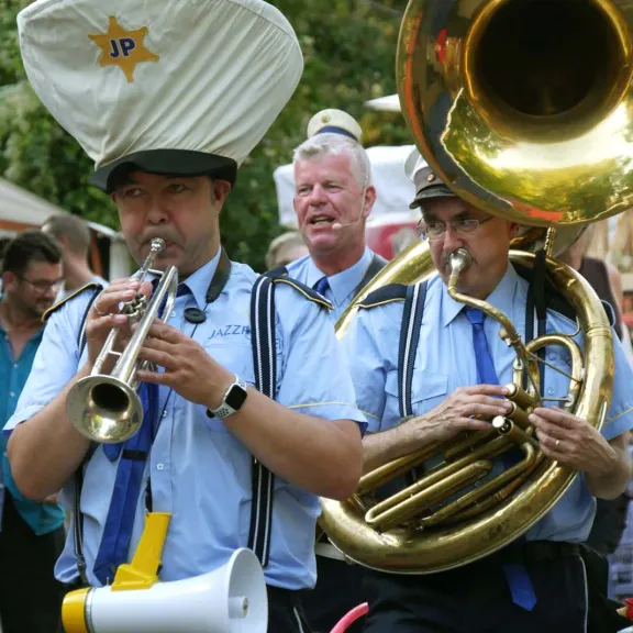 Drei Musiker der Jazz‑Polizei spielen als mobile Marching‑Band auf einem Wein‑ und Winzerfest; zu sehen sind Trompete, Sousaphon und Gesang, umgeben von Marktbesuchern.