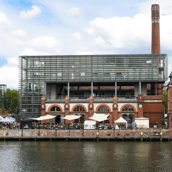 Backstein-Gebäude mit Glasfassade und Schornstein am Fluss, davor Menschen und Zelte.