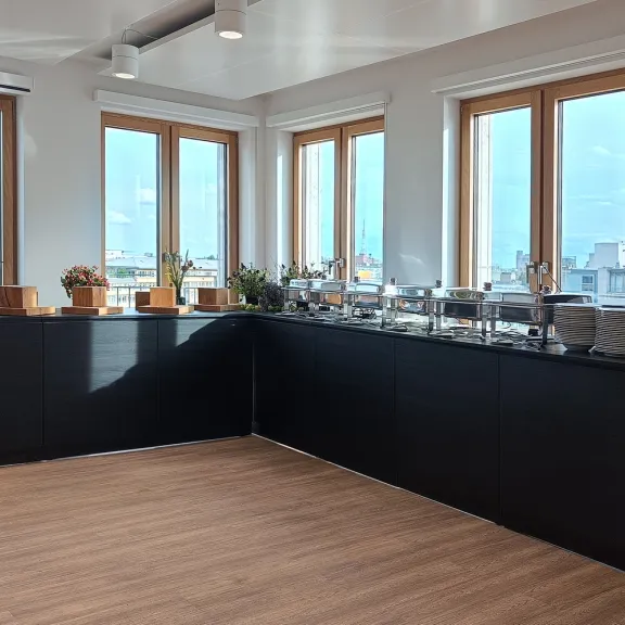 Heller Raum mit Holzboden, Fensterfront und schwarzer Theke, darauf Pflanzen und ein Cateringaufbau.