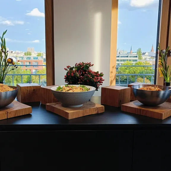 Buffet mit Schüsseln voller Salate vor Fenster mit Ausblick auf Stadt und blauen Himmel.