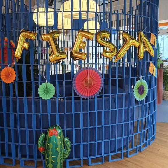 Goldene "FIESTA"-Luftballons und bunte Deko vor blauem Gitter, unten ein Kaktus.