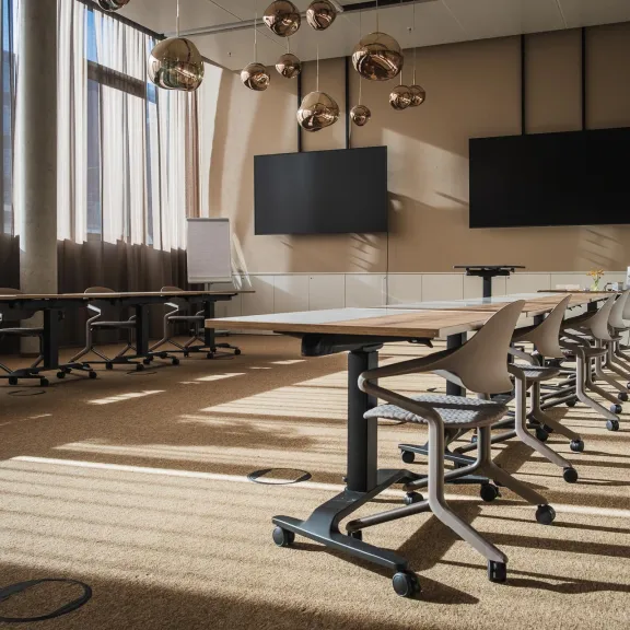 Heller Konferenzraum mit langen Tischen, Stühlen und zwei großen Bildschirmen.