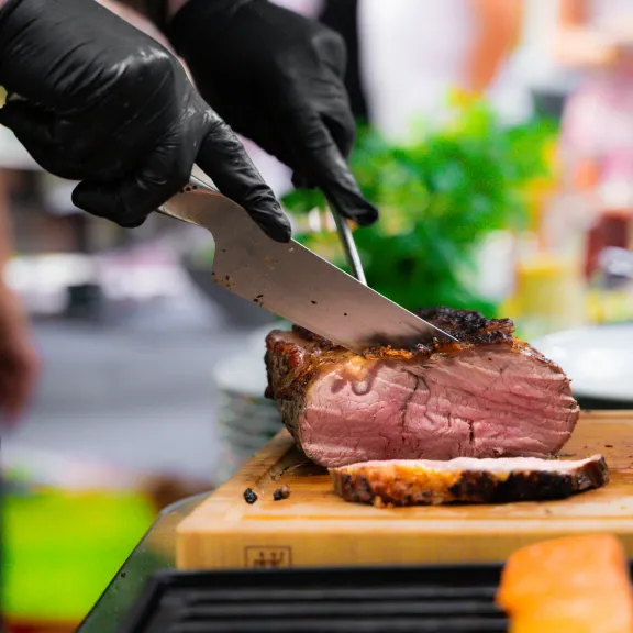 Koch schneidet gebratenes Fleisch auf einem Holzbrett, im Hintergrund Kräuter.