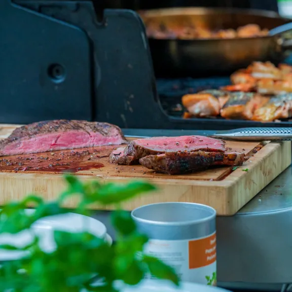 Gegrilltes Steak und Fleisch auf Holzbrett neben Grill mit Gemüse im Hintergrund.