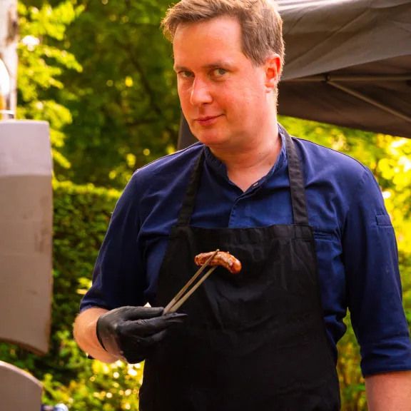 Mann am Grill hält mit einer Zange eine Bratwurst, sommerliche Gartenkulisse.