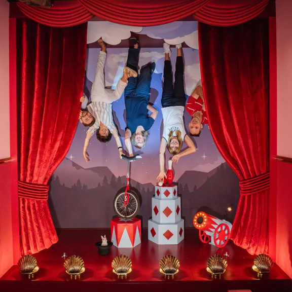 Drei Personen hängen kopfüber auf einer Zirkusbühne mit roten Vorhängen.