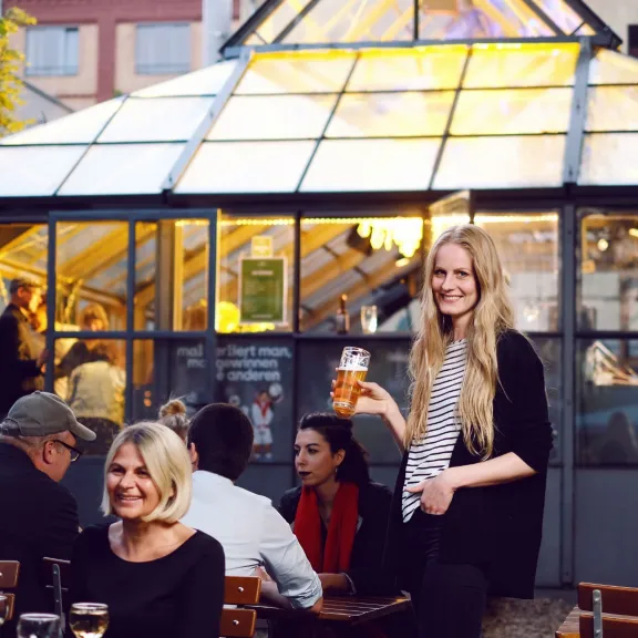 Junge Frau serviert Getränke im sonnigen Außenbereich eines Restaurants mit Glasfassade.