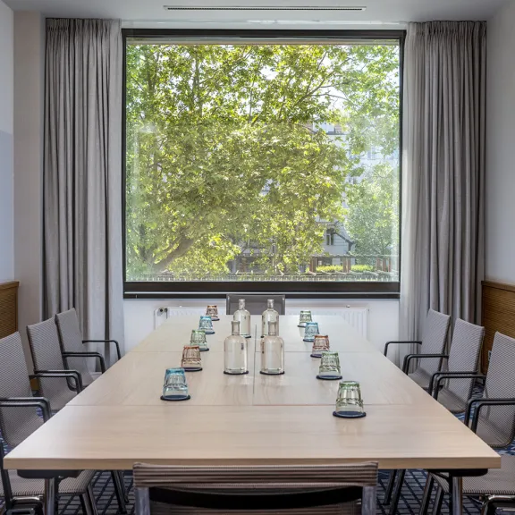 Moderner Konferenzraum mit großem Tisch, Stühlen und Blick auf grüne Bäume durchs Fenster.
