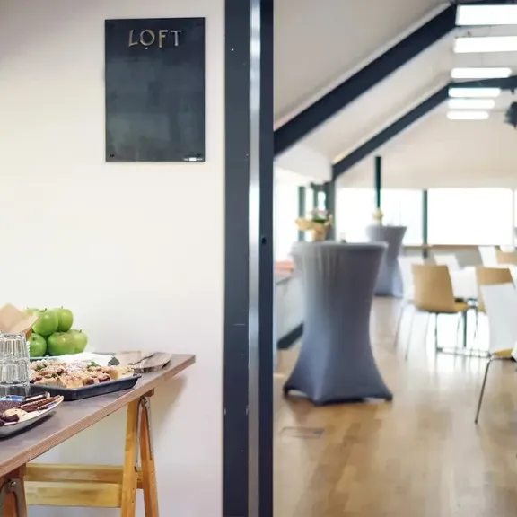 Heller Veranstaltungsraum mit gedeckten Tischen und Buffet im Vordergrund.