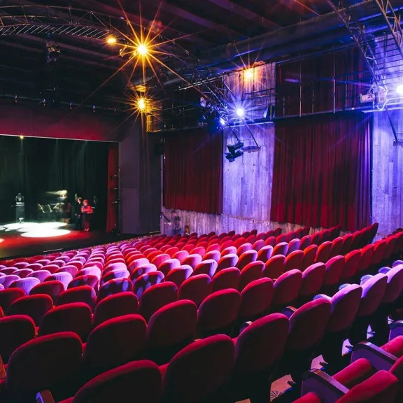 Leerer Theatersaal mit roten Sitzen, Bühne beleuchtet, violette und blaue Scheinwerfer.