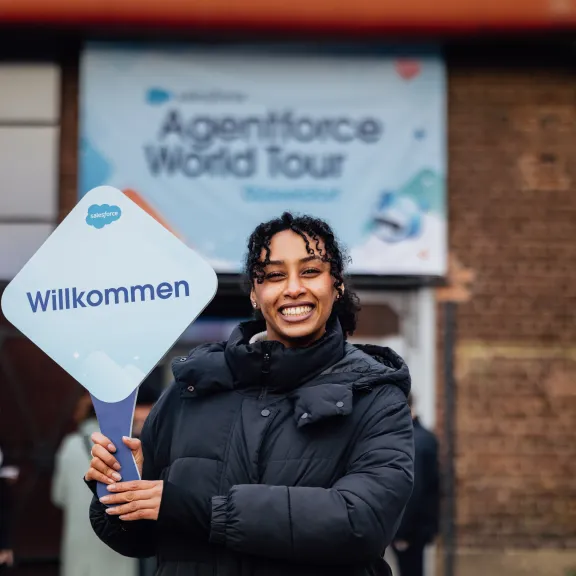 Transferhostess von cinnamon in schwarzer Winterjacke begrüßt Gäste lächelnd mit einem Willkommensschild bei der Ankunft.