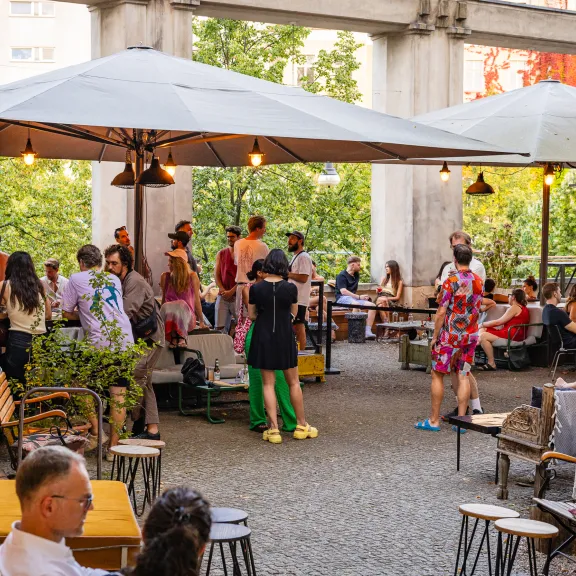 Menschen sitzen und stehen in einem belebten Café-Garten unter Sonnenschirmen.