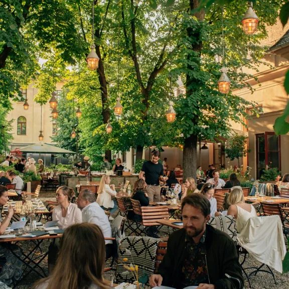 Menschen sitzen und essen entspannt im schattigen Gartenrestaurant unter grünen Bäumen.