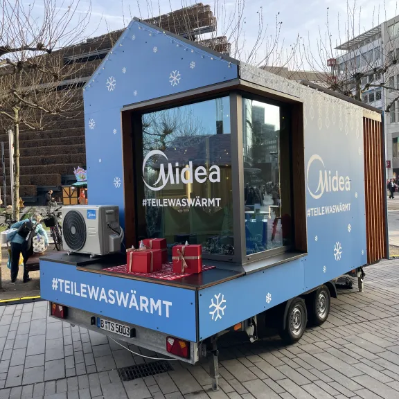 Blauer Anhänger in Hausform mit Glasfront, Heizung, Geschenkpaketen und Stadt im Hintergrund.