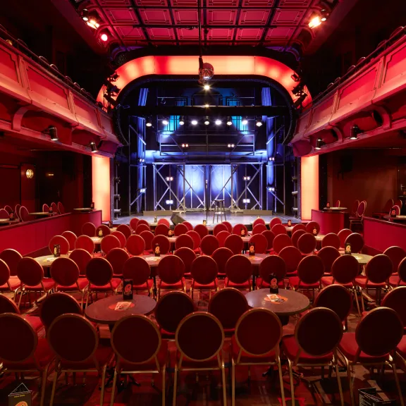 Rot beleuchteter Theatersaal mit runden Tischen und Stühlen, Blick auf die Bühne.