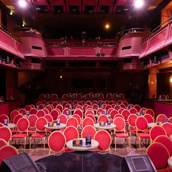 Leerer Theatersaal mit roten Stühlen und runden Tischen, Bühne im Vordergrund.
