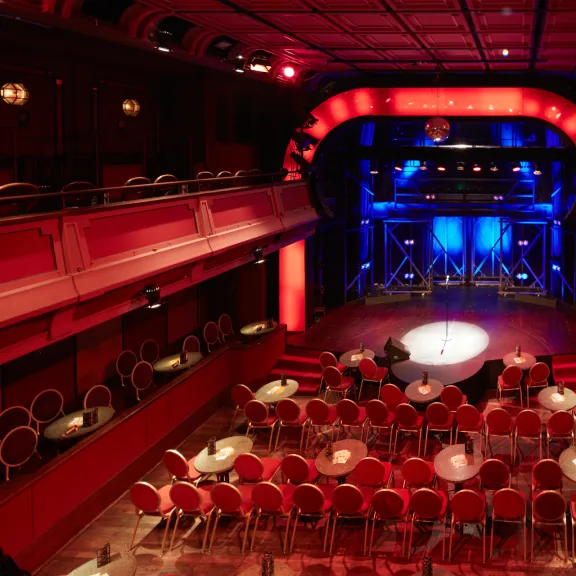 Roter Theatersaal mit runden Tischen, leeren Stühlen und beleuchteter Bühne.
