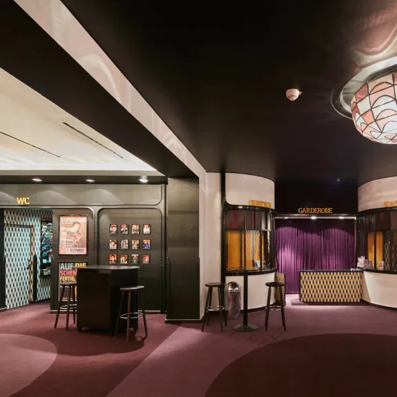 Modernes Kino-Foyer mit lila Teppich, großer Deckenlampe und Kinoplakaten an den Wänden.