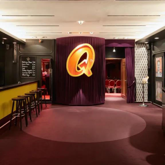 Foyer eines Theaters mit rotem Teppich und großem gelbem Q an violettem Vorhang.