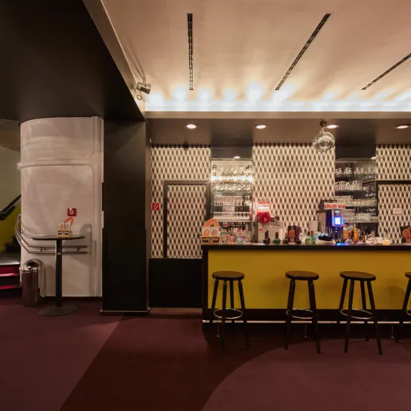 Moderne Bar mit gelber Theke, dunklen Hockern und gemusterten Wänden, warme Beleuchtung.