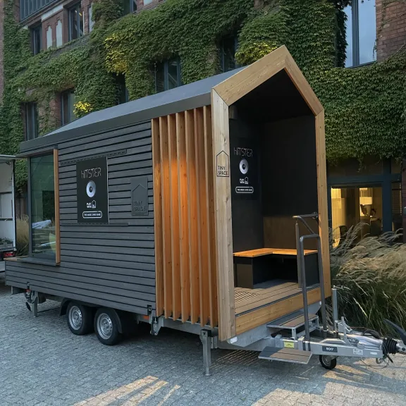 Modernes Tiny House auf einem Anhänger, umgeben von begrünten Gebäuden.