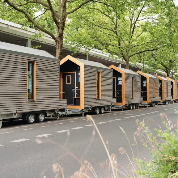 Reihe moderner Tiny Houses auf Anhängern unter Bäumen an einer Straße.