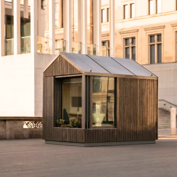 Kleines, modernes Holzhaus mit großen Fenstern vor städtischem Gebäude.