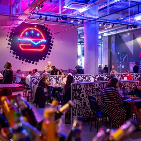 Modernes Restaurant mit vielen Gästen, bunte Beleuchtung und stilvollem Burger-Logo an der Wand.