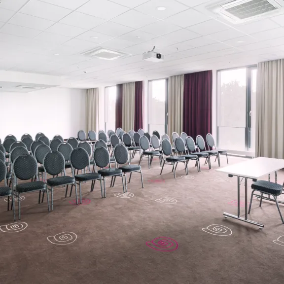 Leerer Konferenzraum mit Stuhlreihen und einem Tisch, große Fenster mit Vorhängen.