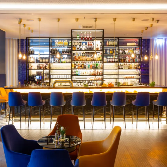 Moderne Bar mit beleuchtetem Tresen, blauen Stühlen und bunten Sesseln im Vordergrund.