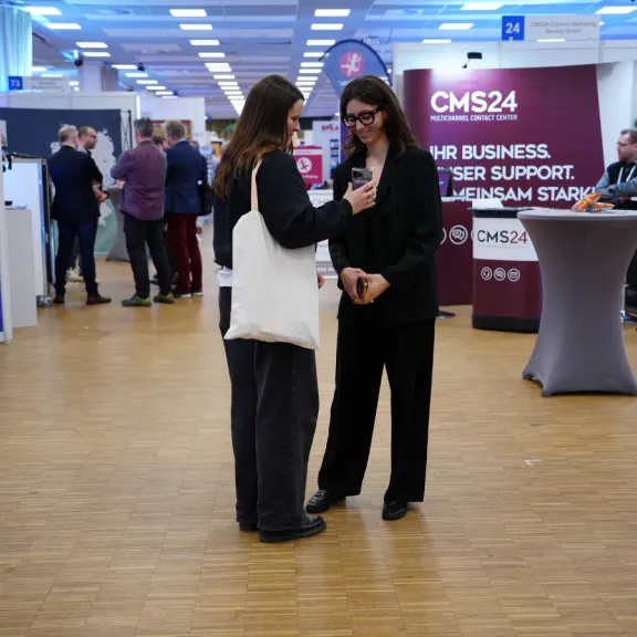 Zwei Frauen unterhalten sich auf einer Messe mit Infoständen im Hintergrund.