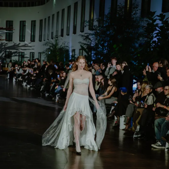Model in weißem Kleid läuft auf einem beleuchteten Laufsteg, Zuschauer sitzen am Rand.