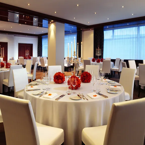 Elegant gedeckter Bankettsaal mit weißen Stühlen und runden Tischen, rote Blumendekoration.