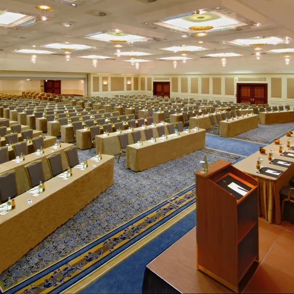 Großer Konferenzsaal mit vielen leeren Tischen und Stühlen, Rednerpult im Vordergrund.