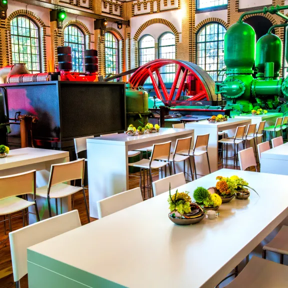 Modern dining tables with floral centerpieces in a colorful industrial hall with large machinery.