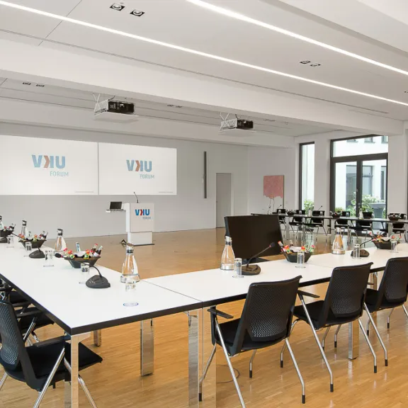 Heller Konferenzraum mit U-förmigen Tischen, Stühlen und Präsentationsbildschirmen.