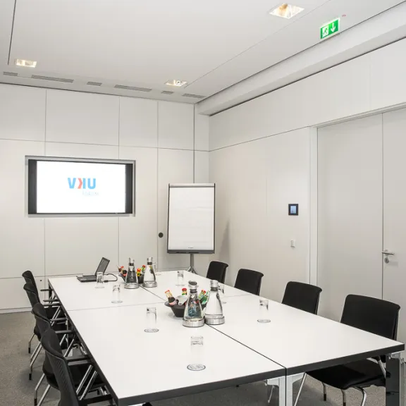 Moderner Konferenzraum mit großem Tisch, schwarzen Stühlen und Präsentationsbildschirm.