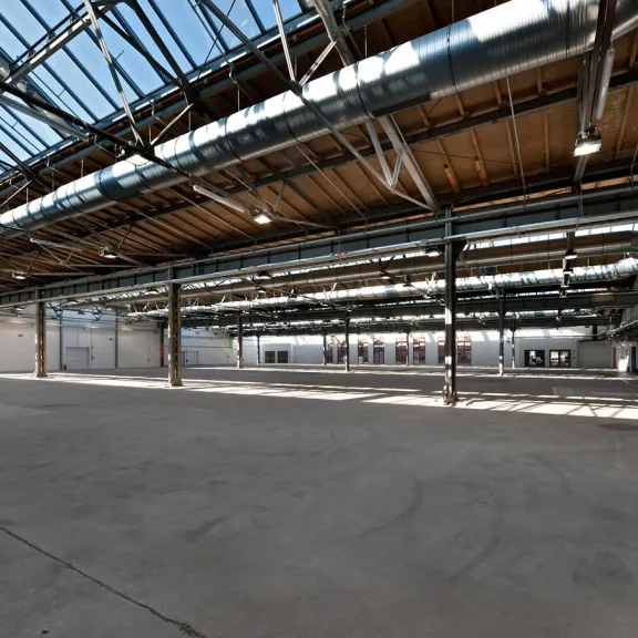 Große, leere Industriehalle mit Glasdach und sichtbaren Metallträgern.