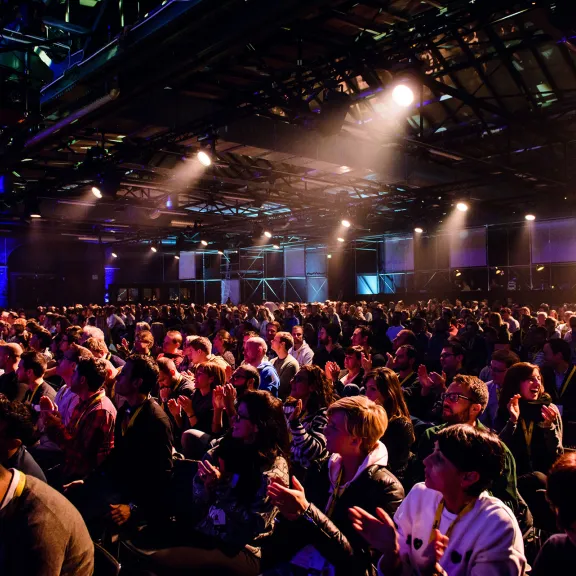 Großes Publikum sitzt in einem dunklen, beleuchteten Konferenzsaal.