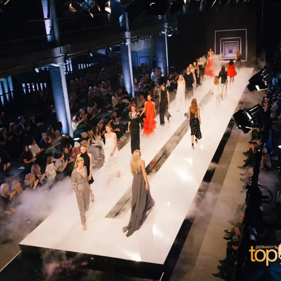 Models laufen auf einem beleuchteten Laufsteg, umgeben von Publikum und Nebel.