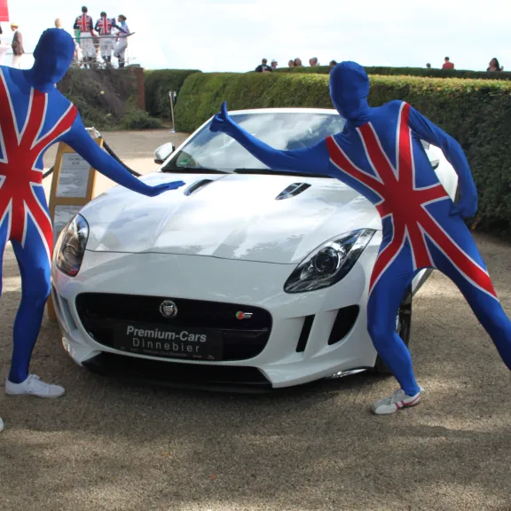 Zwei Personen im Union-Jack-Anzug posieren vor einem weißen Jaguar-Sportwagen.