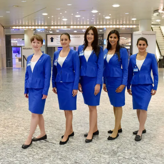 Fünf Flugbegleiterinnen in blauen Uniformen stehen in einer hellen Flughafenhalle.