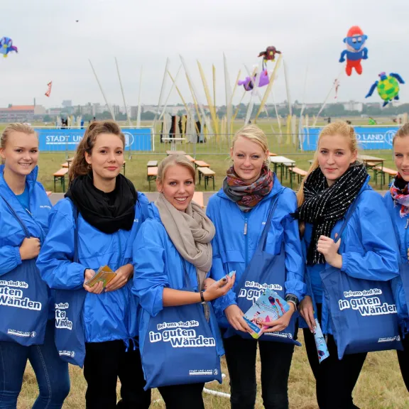 Sieben junge Frauen in blauen Jacken stehen draußen vor Bänken und bunten Drachen.