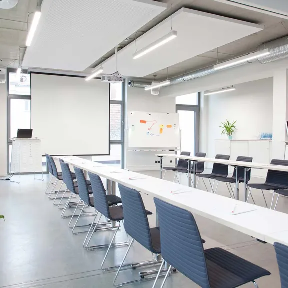 Heller, moderner Konferenzraum mit langen Tischen, Stühlen und großer Projektionsfläche.