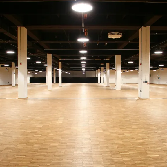 Große, leere Halle mit Holzfußboden und weißen Säulen unter Deckenlampen.
