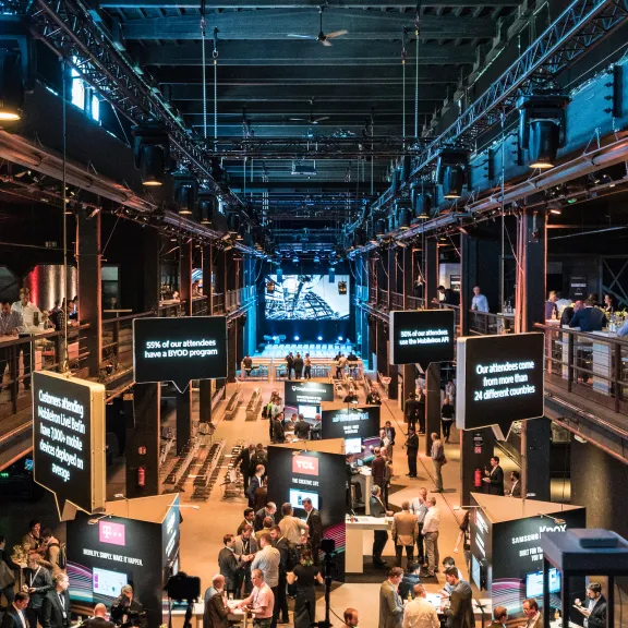 Große Messehalle mit Ständen, Besuchern und einer beleuchteten Bühne im Hintergrund.