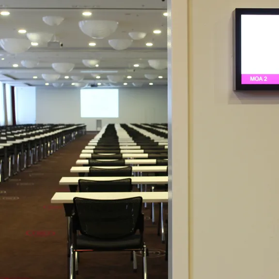 Leerer Konferenzraum mit Reihen von Tischen und Stühlen, Bildschirm an der Wand.